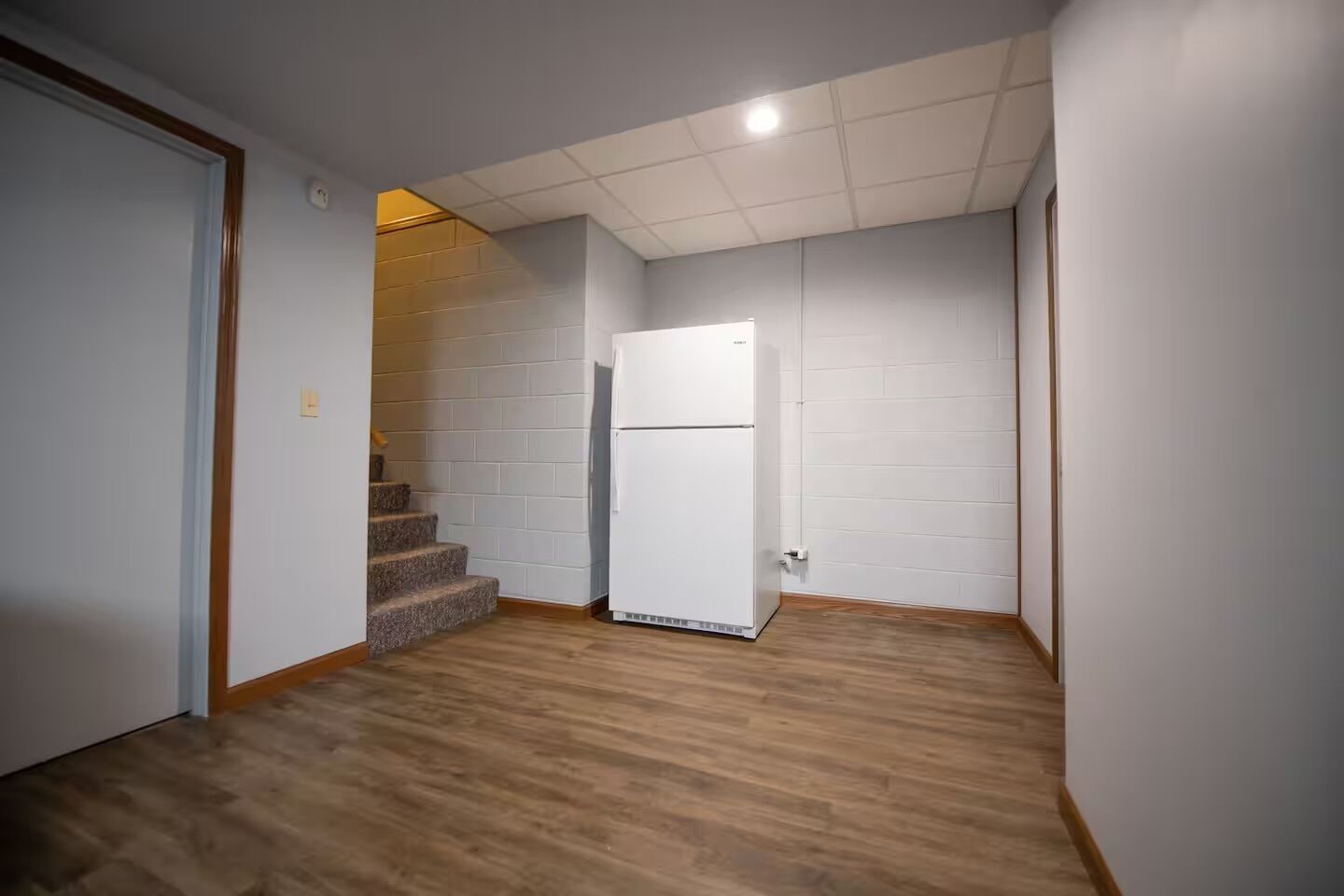 A room with a white refrigerator, a wooden floor, and a white brick wall.