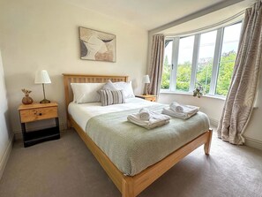 A neatly made bed with a patterned bedspread, a bedside table with a lamp, a nightstand with a lamp, and a window with curtains. - 3-BR Bungalow w/ Spacious Living & Private Garden (Lincolnshire)