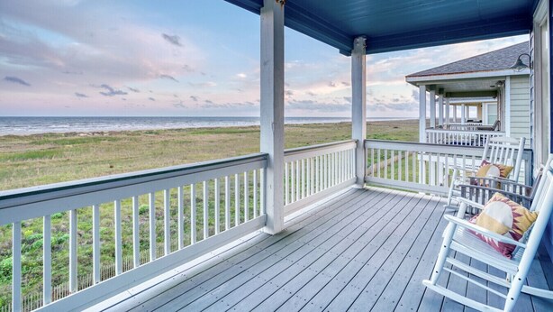 A covered porch with a view of the beach, white railings, and a seating area with a chair and a small table.