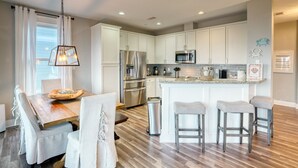 A modern kitchen with white cabinets, stainless steel appliances, a wooden dining table, and a bar area with stools.