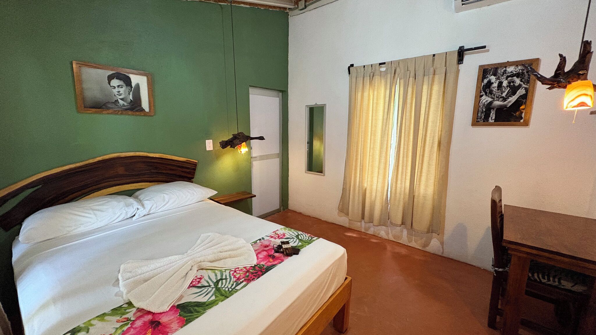Un dormitorio con cama, una silla, una mesita de noche, una lámpara de pared, un cuadro enmarcado y una ventana con cortinas.