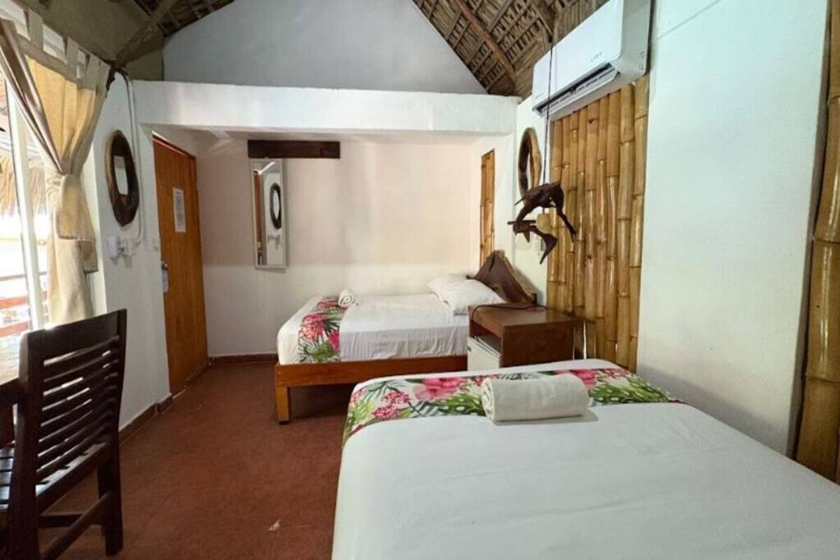 Una habitación con dos camas, una silla de madera, una mesita y una lámpara en la pared.