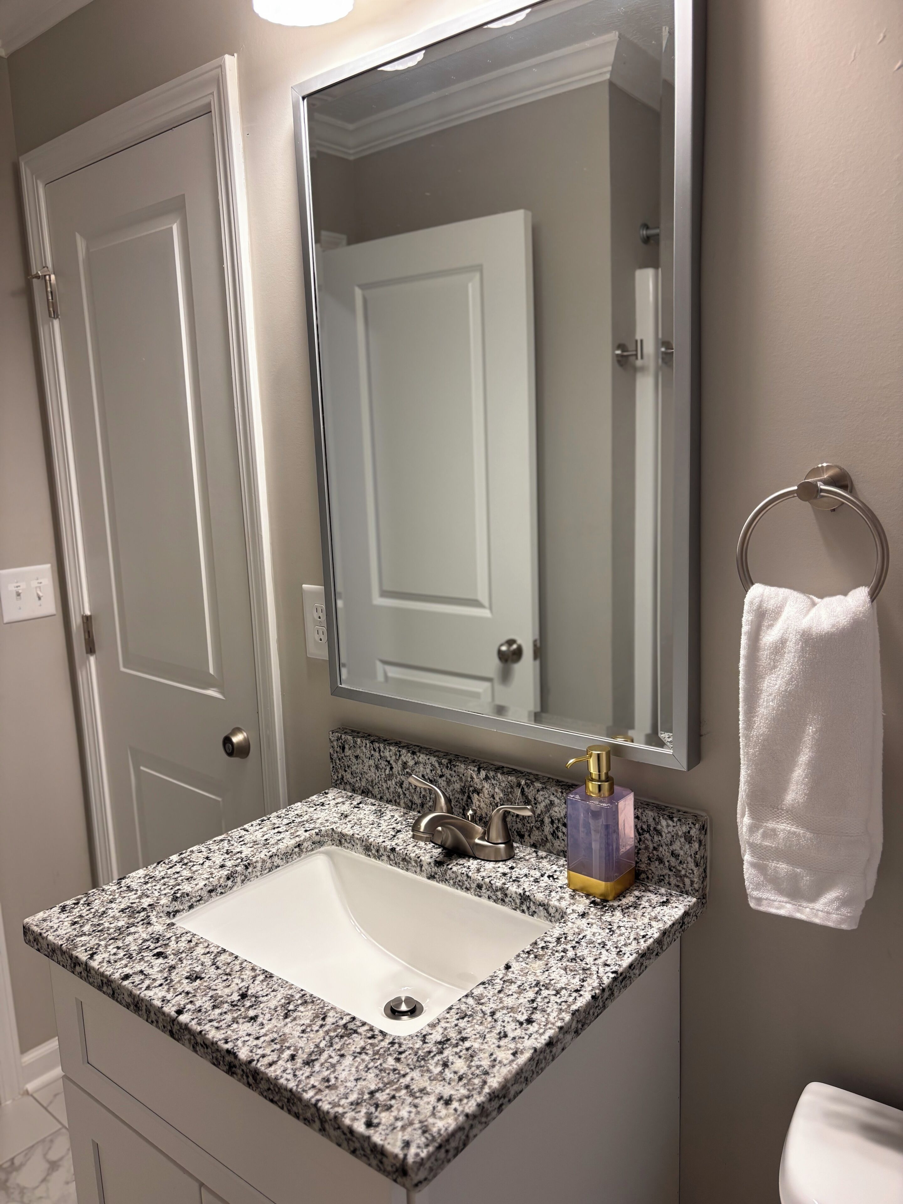 Un baño con lavamanos de encimera de granito, un espejo, un tozalengua y una puerta.