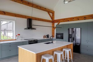 A modern kitchen with a large island, wooden beams, a stainless steel refrigerator, and a window with a view. - Black Ridge Barn - Minimalist Restored Barn (Prince Edward County)