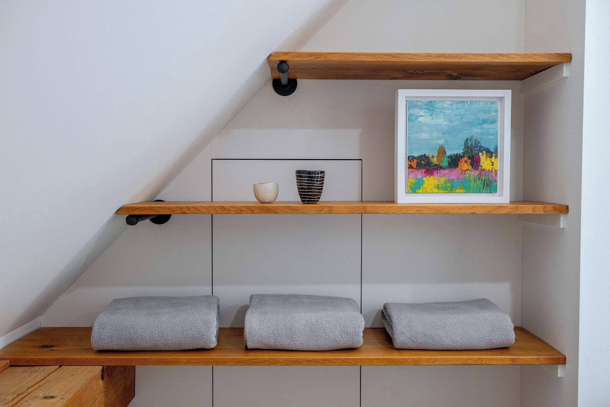 Une pièce avec une étagère en bois, un tableau encadré, un petit bol et trois serviettes pliées.