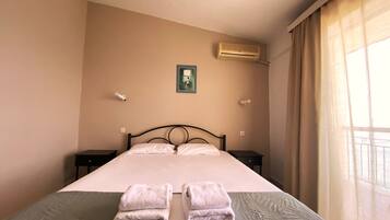 A hotel room with a bed, two bedside tables, an air conditioning unit, a window with curtains, and a small framed picture on the wall.