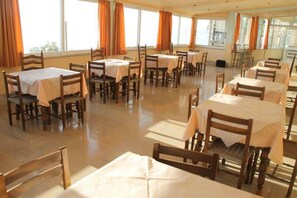 A dining area with tables and chairs, large windows with curtains, and a polished floor. - El Greco Hotel (Corfu)