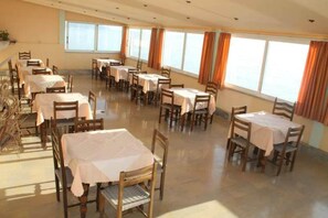 A dining area with multiple tables and chairs, large windows with curtains, and a tiled floor. - El Greco Hotel (Corfu)