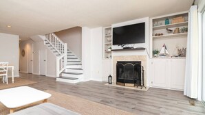 Un salon moderne doté d’une cheminée, d’un téléviseur à écran plat fixé au-dessus, d’un escalier menant à une autre pièce et d’un coin repas avec une table et des chaises.