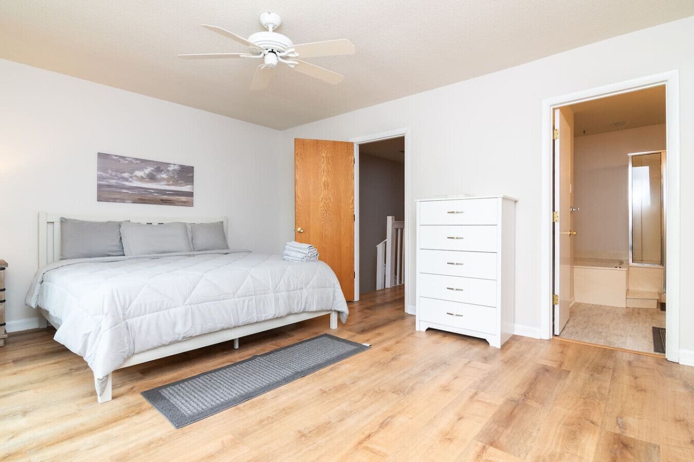 A bedroom with a bed, a nightstand, a dresser, a ceiling fan, a wooden door, and a bathroom visible through an open door.