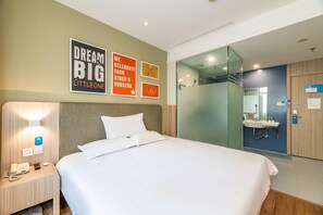Habitación de hotel con cama, mesita de noche con teléfono y control remoto, pared decorada con carteles motivacionales y baño con mampara de vidrio.