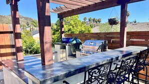 Uma área de bar externa com churrasqueira, banquetas e uma estrutura de pérgola.