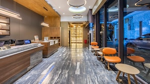 A modern hotel lobby with a reception desk, seating area, and a view of the street through large windows.