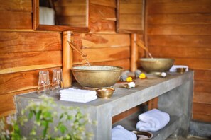 Interior de una cabaña de madera con un banco de piedra, un cuenco grande de latón y un cuenco más pequeño en la encimera.