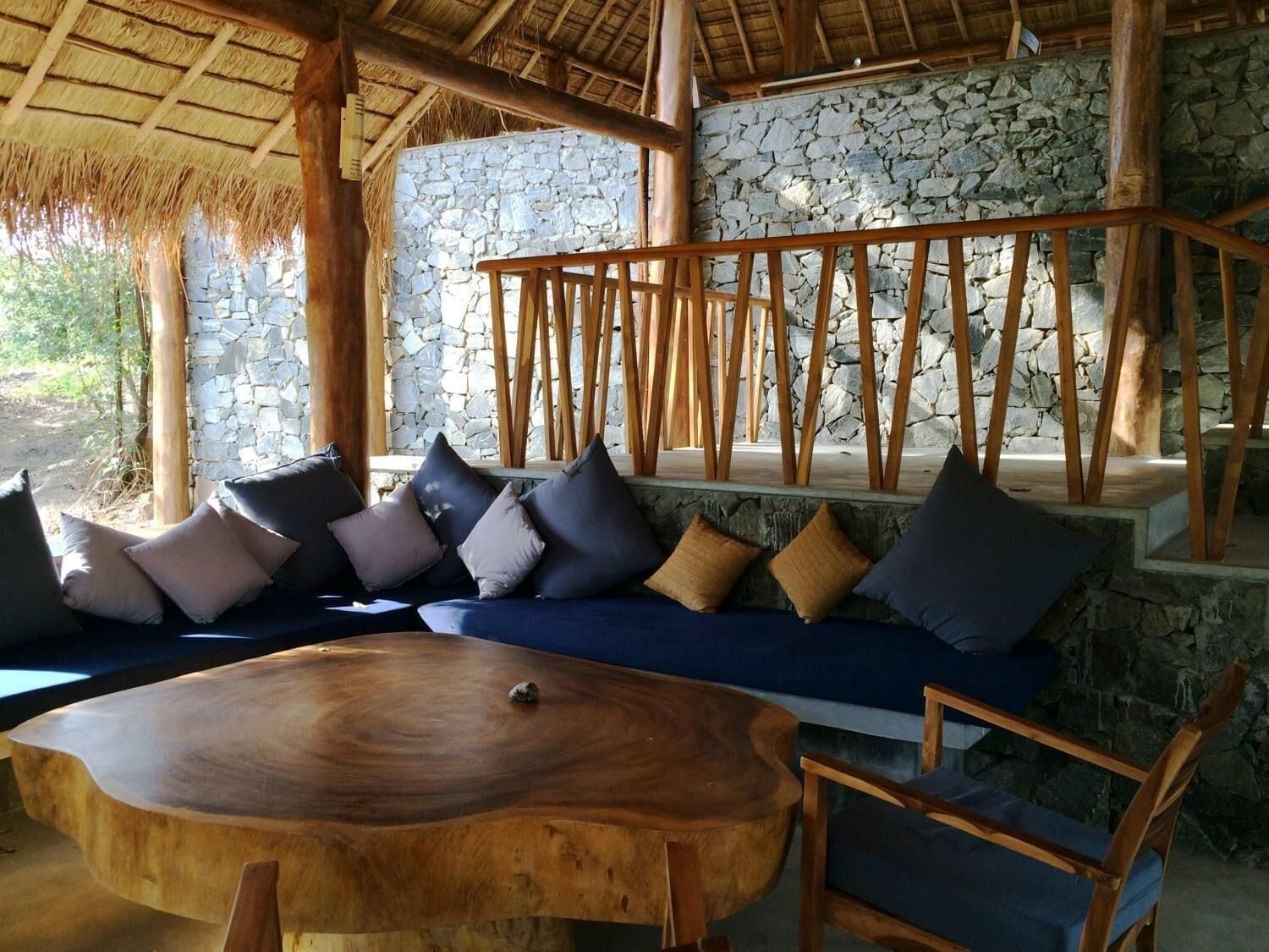 A wooden bench with cushions, a round wooden table, and a stone wall in a rustic setting.