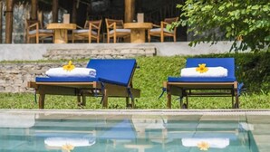 A poolside area with a thatched-roof structure, wooden lounge chairs, and a clear pool.