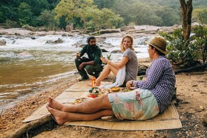 Three people sitting on a mat near a river with food and drinks, surrounded by rocks and greenery. - Gal Oya Lodge (MADULLA)