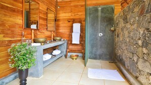 A wooden bathroom with a wooden bench, a mirror, a sink with bowls, a towel rack, and a stone wall.