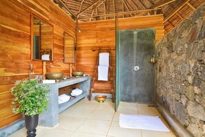 Un baño de madera con banca, espejo, lavamanos con recipientes, toallero y pared de piedra.