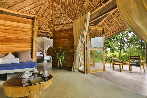 A wooden cabin with a thatched roof, a sofa, a table with a teapot, and a view of greenery outside.