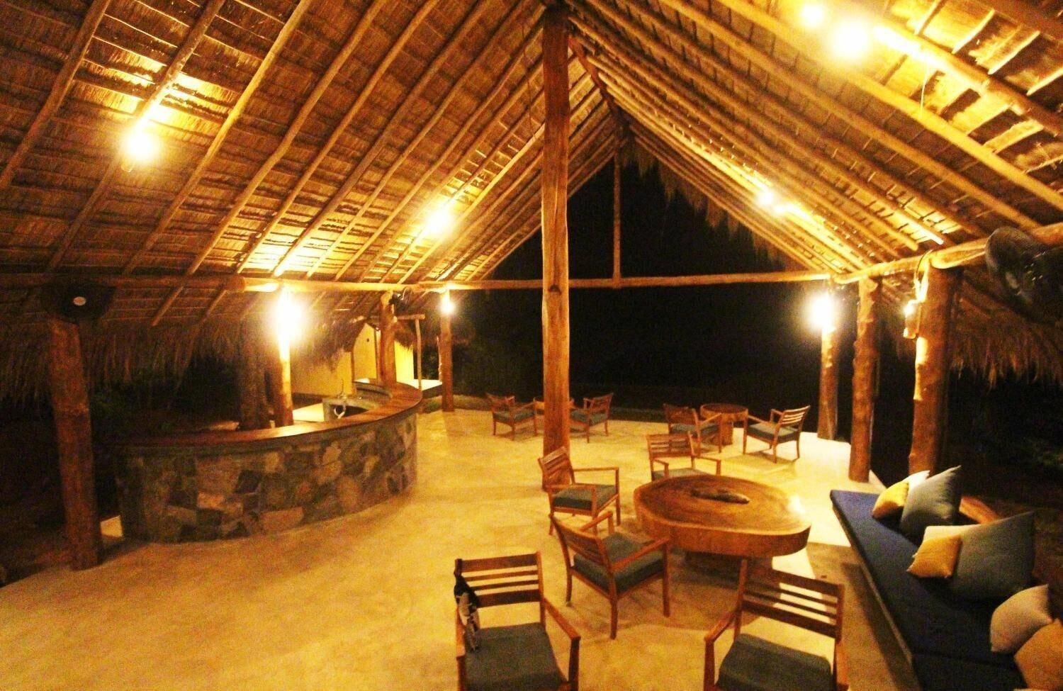 An outdoor lounge area with a stone bar, wooden furniture, and a thatched roof structure.