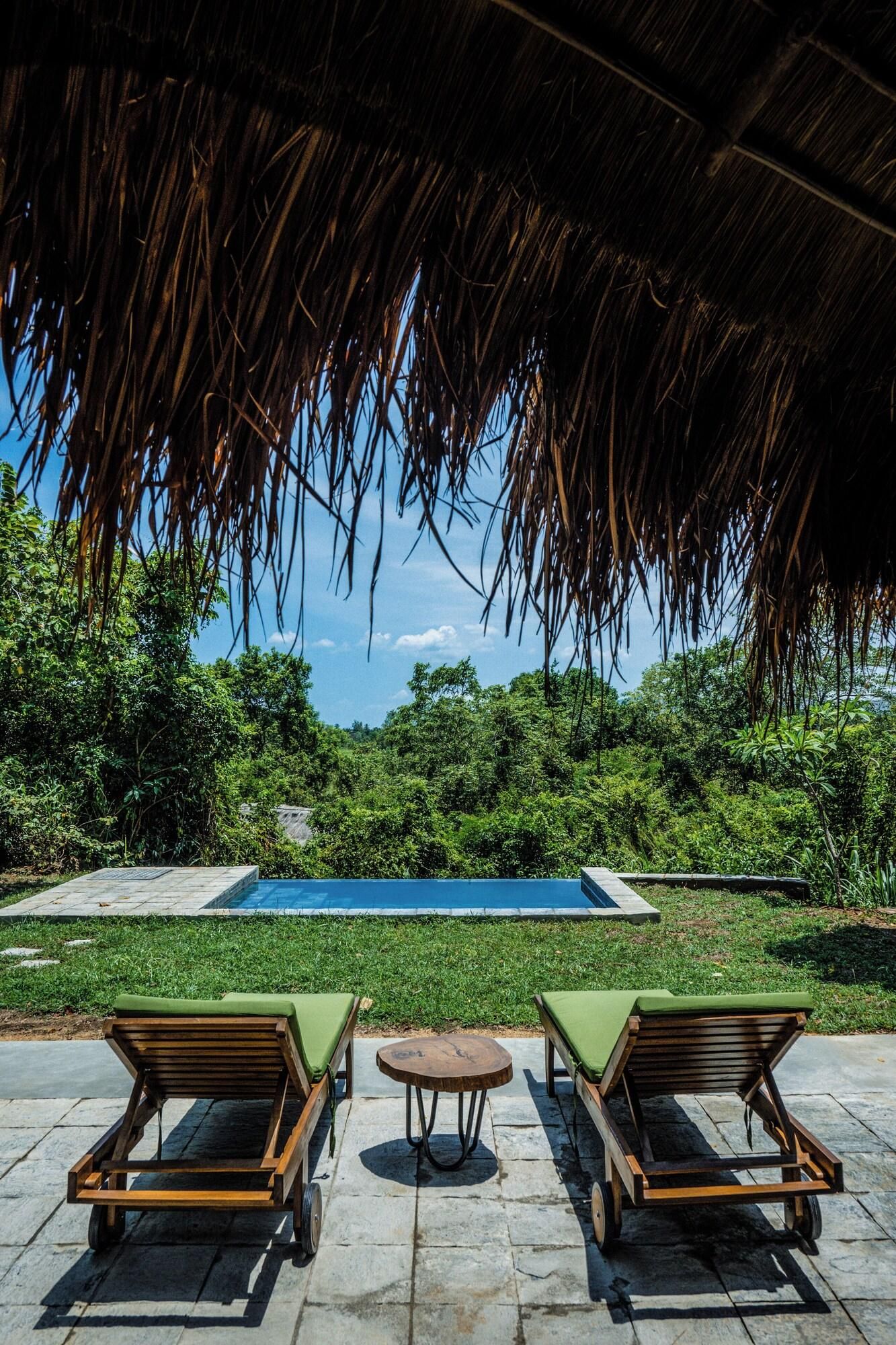 Un espace aménagé au bord de la piscine, doté d’un toit de chaume, de deux chaises longues en bois, d’une petite table ronde et offrant une vue dégagée sur une piscine entourée d’une végétation luxuriante.