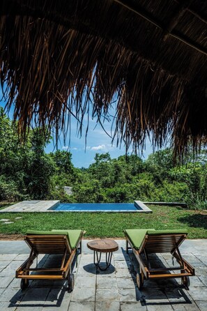 A poolside area with a thatched roof, two wooden lounge chairs, a small round table, and a clear view of a swimming pool surrounded by lush greenery. - Gal Oya Lodge (MADULLA)