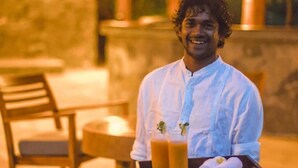 A person holding a tray with drinks and towels, standing in an outdoor area with a thatched roof and seating. - Gal Oya Lodge (MADULLA)