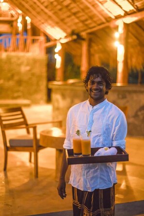 A person holding a tray with drinks and towels, standing in an outdoor area with a thatched roof and seating. - Gal Oya Lodge (MADULLA)