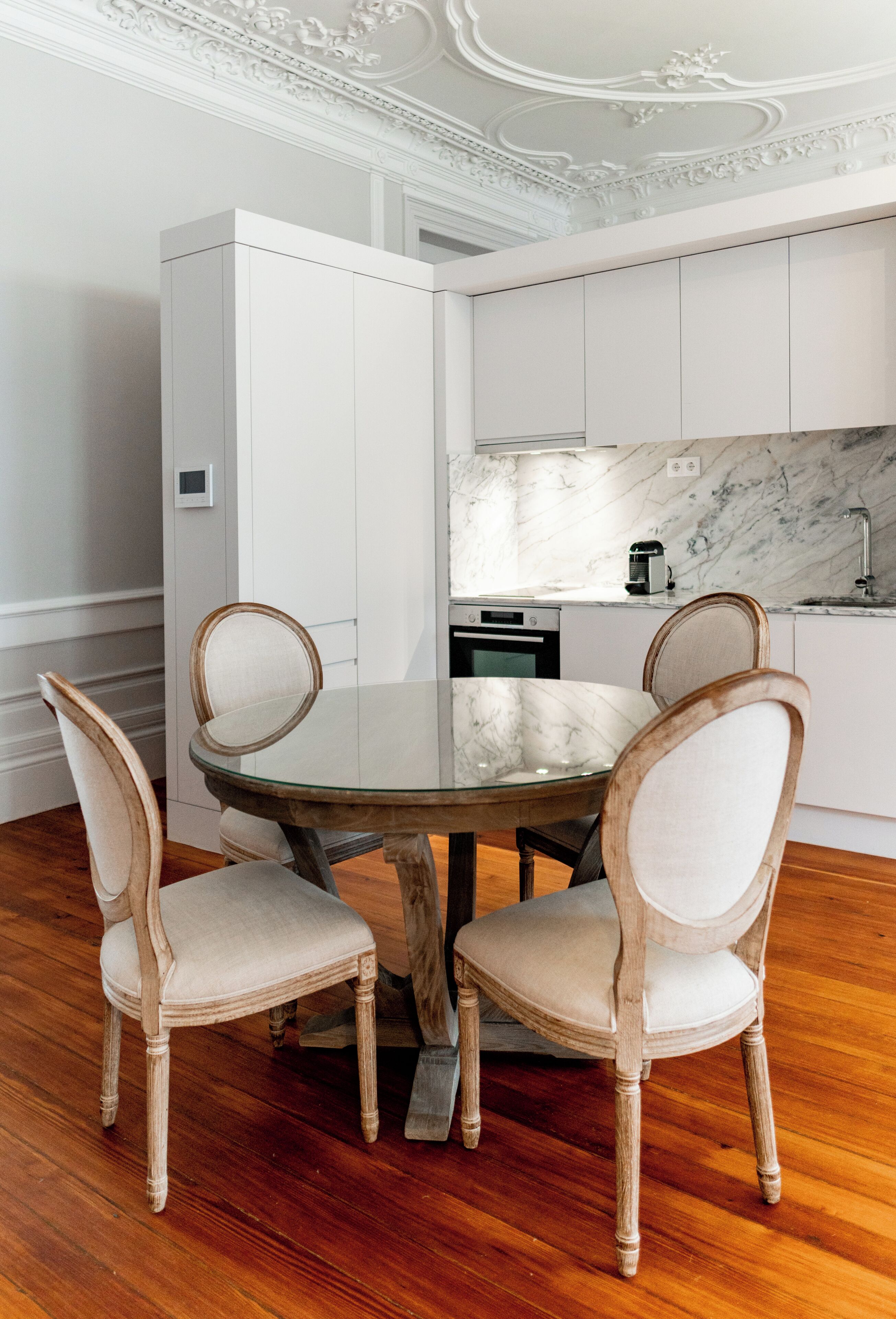 A dining area with a round table and four chairs, a kitchen with white cabinets and marble backsplash, and wooden flooring.