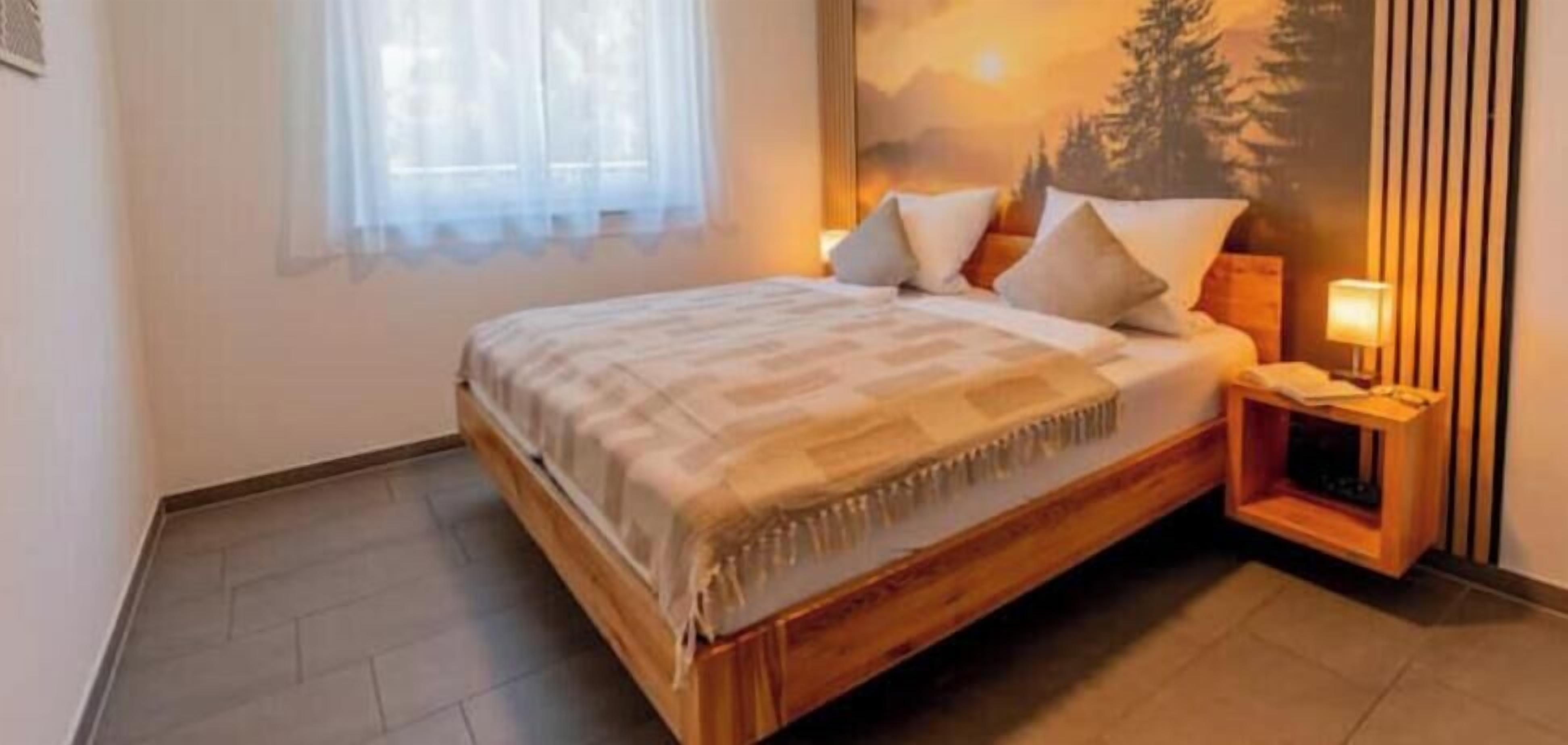 A bedroom with a wooden bed, a nightstand, a lamp, and a window with sheer curtains.
