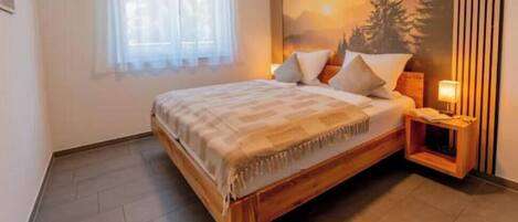 A bedroom with a wooden bed, a nightstand, a lamp, and a window with sheer curtains.