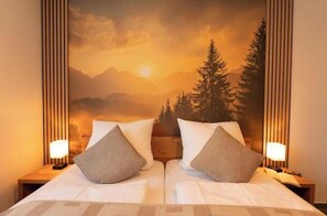 Ein Doppelbett mit zwei Kissen, ein Nachttisch mit Lampe sowie ein Wandbild mit Berglandschaft.