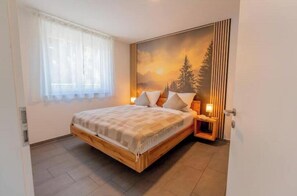 A bedroom with a bed, a nightstand, a lamp, a window with curtains, and a wall with a painting. - 2-Bedroom Apartment with Living Room and Garden (Titisee-Neustadt)