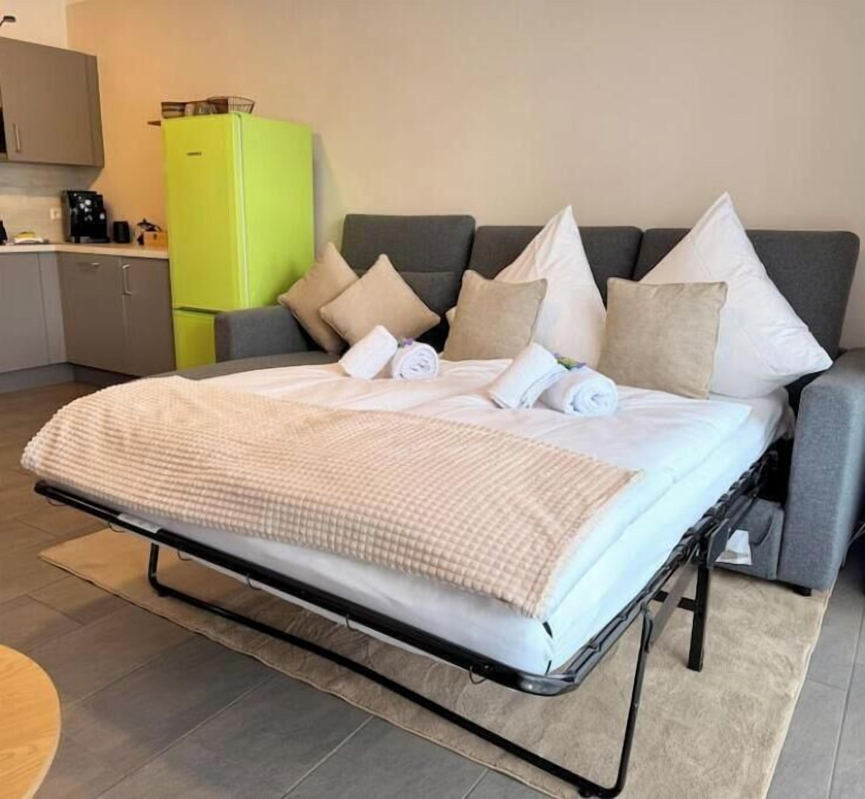 A bed with a checkered blanket, grey sofa with multiple pillows, a green refrigerator, and a kitchen area with cabinets and a coffee machine.