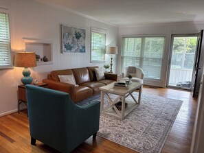 Un salon avec un canapé en cuir marron, un fauteuil turquoise et une table basse. On y trouve deux lampes, un tableau et une vue à travers la porte vitrée.