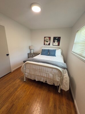 A bedroom with a bed, a nightstand, a lamp, two wall art pieces, and a window with blinds.