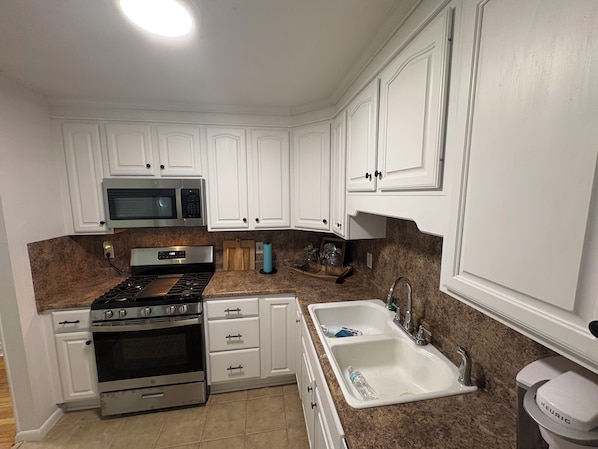 Una cocina con gabinetes blancos, microondas, estufa, lavabo y cafetera Keurig.