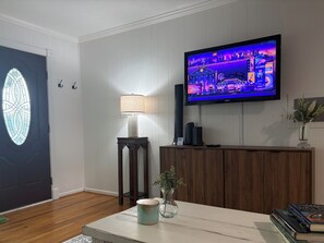Ett vardagsrum med en platt-TV som är monterad på väggen, ett träbord med en vas och böcker, en lampa på ett sidobord och en dörr med ett fönster av färgat glas.