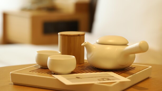 A tea set on a tray with a wooden mat, a bed, and a nightstand in the background. - Ji Hotel (Chengdu)