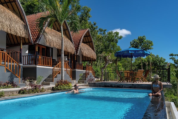 A swimming pool with a thatched-roof building, palm trees, and a clear blue sky. - Sunrise Penida Hill by ABM (Penida Island)