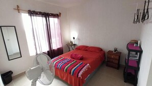 Une chambre à coucher comprenant un lit, un ventilateur sur pied, une table de chevet, une commode et un miroir.