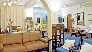 A living room with a brown sofa, blue chairs, a wooden chair, and a painting on easel. - Lassion Beach Hotel (Sitia)