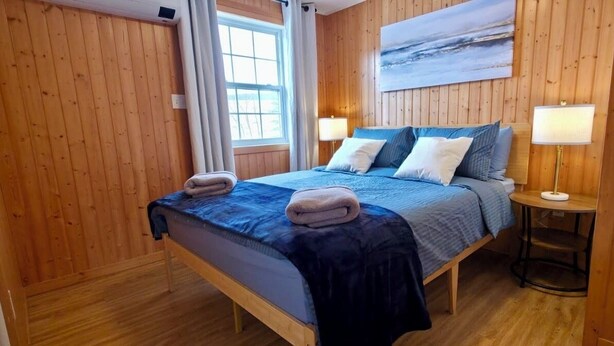 Una habitación con paneles de madera, una cama, dos lámparas de noche, un ventanal y un cuadro en la pared.