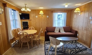 Huoneessa on puupanelilla verhoiltu seinä, sohva, sohvapöytä, ruokapöytä tuoleineen, takka ja televisio.