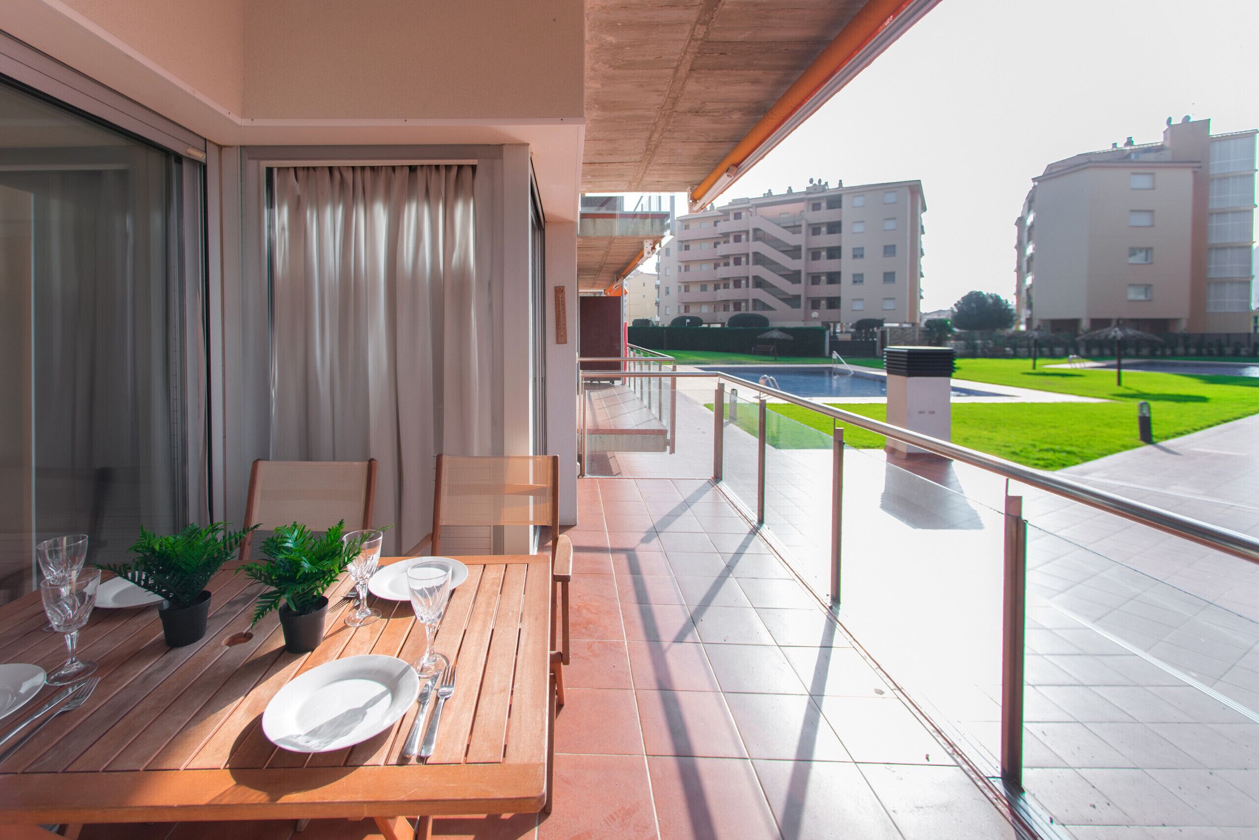 A balcony with a wooden table set for dining, a view of a green lawn, and buildings in the background.