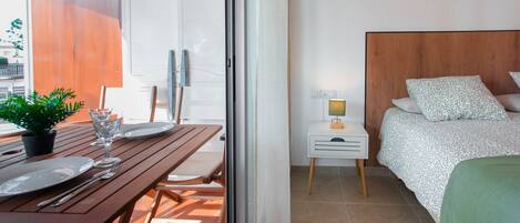 A room with a bed, a bedside table with a lamp, a window, and a balcony with a table set for two.