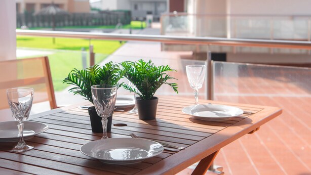 Et træbord med tallerkener, glas og krukker med planter på en balkon med udsigt til bygninger og et grønt område.