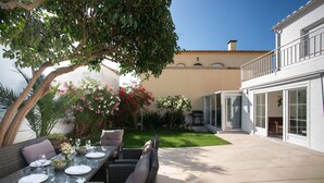 Eine Terrasse im Freien mit Korbmöbeln, einem Esstisch für vier Personen und einem Garten mit verschiedenen Pflanzen und Blumen.