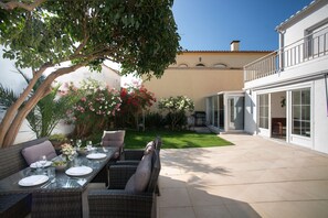 Eine Terrasse im Freien mit Korbmöbeln, einem Esstisch für vier Personen und einem Garten mit verschiedenen Pflanzen und Blumen.